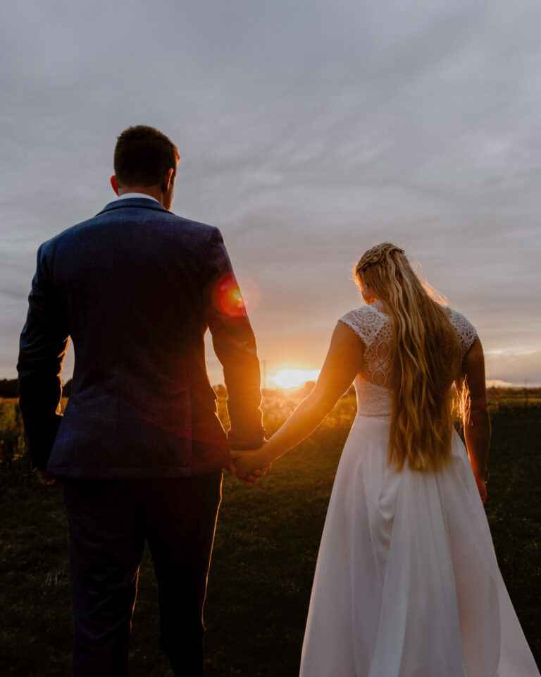 Brautpaar geht bei Sonnenuntergang Hand in Hand durchs Feld – einer der schönsten Momente für Paarfotos zur Hochzeit