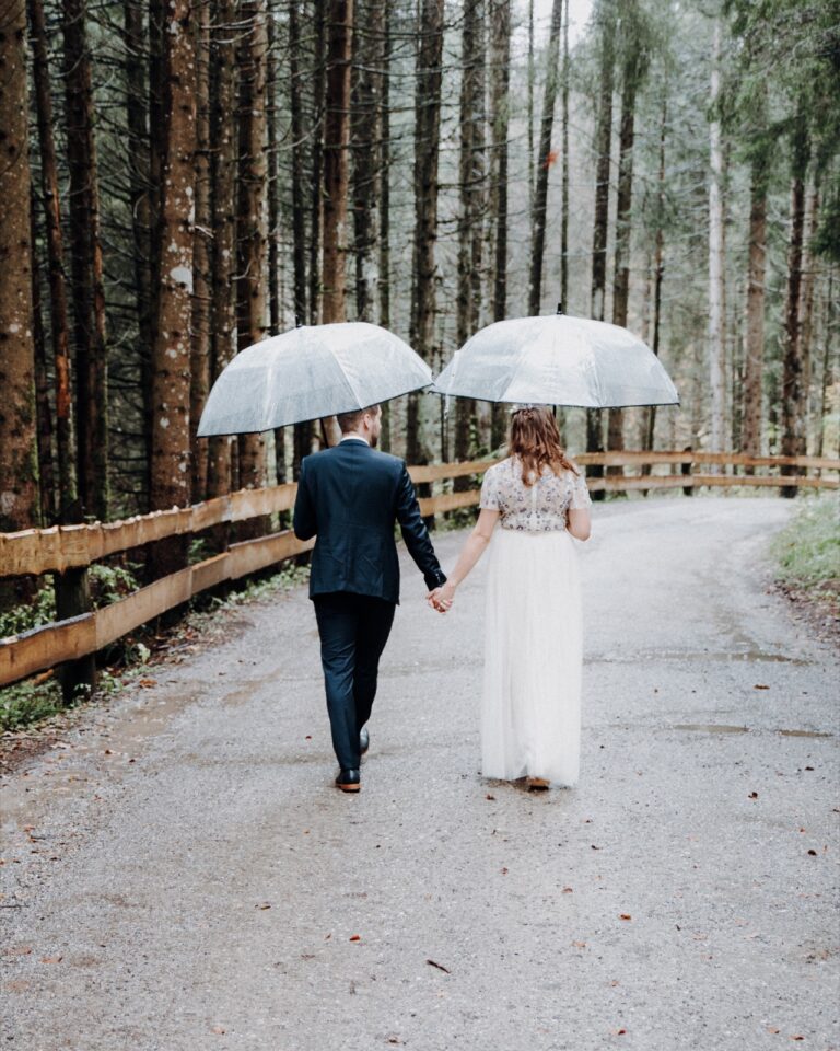 Brautpaar von hinten unter zwei durchsichtigen Regenschirmen auf einem Waldweg, Regentropfen sichtbar