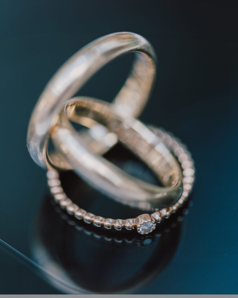 Zwei Eheringe und ein Verlobungsring liegen ineinander verschlungen auf dunklem Glas