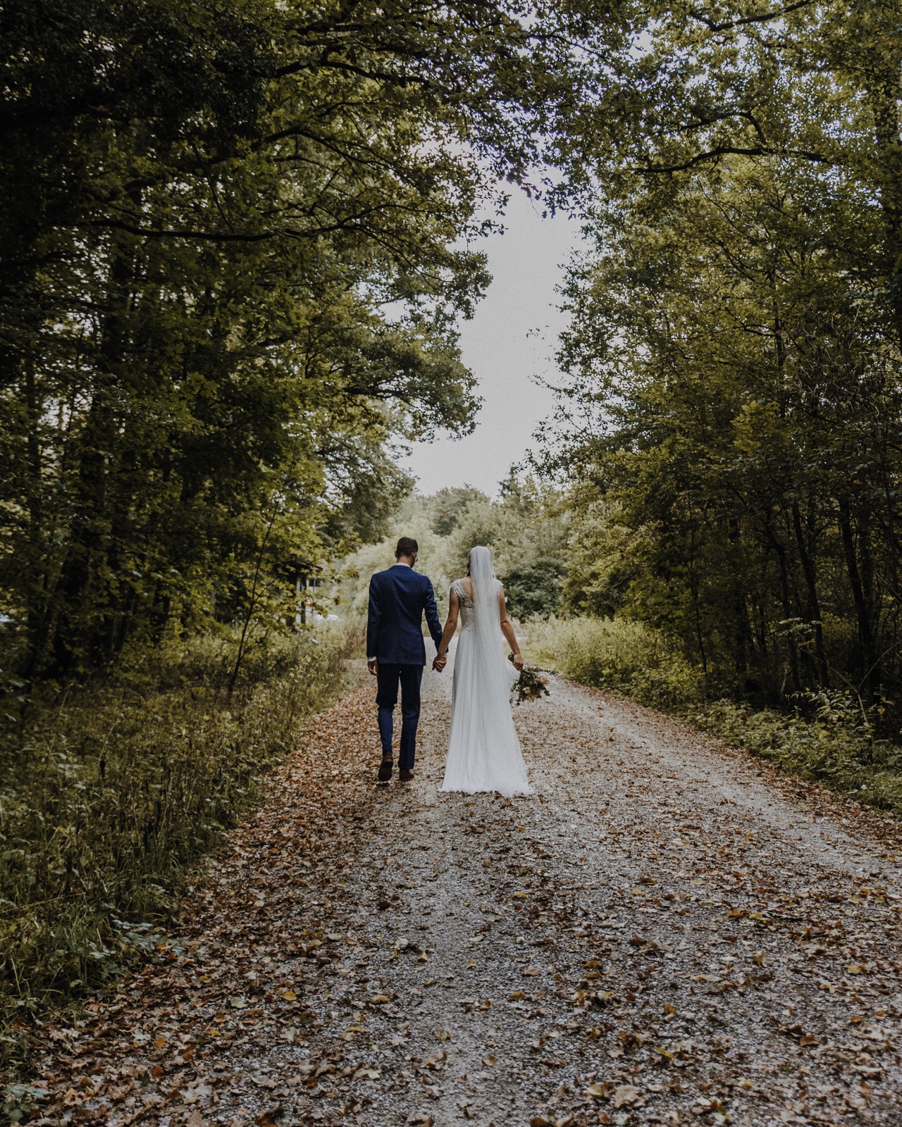 Brautpaar geht Hand in Hand von hinten gesehen einen herbstlichen Waldweg entlang