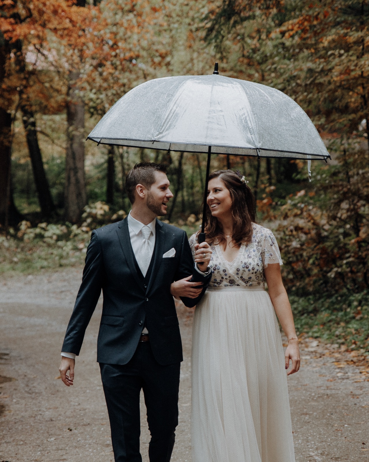 Brautpaar lacht sich unter einem gemeinsamen Regenschirm an, herbstliche Bäume im Hintergrund – ein typisches Beispiel für gelungene Hochzeitsfotos bei Regen.