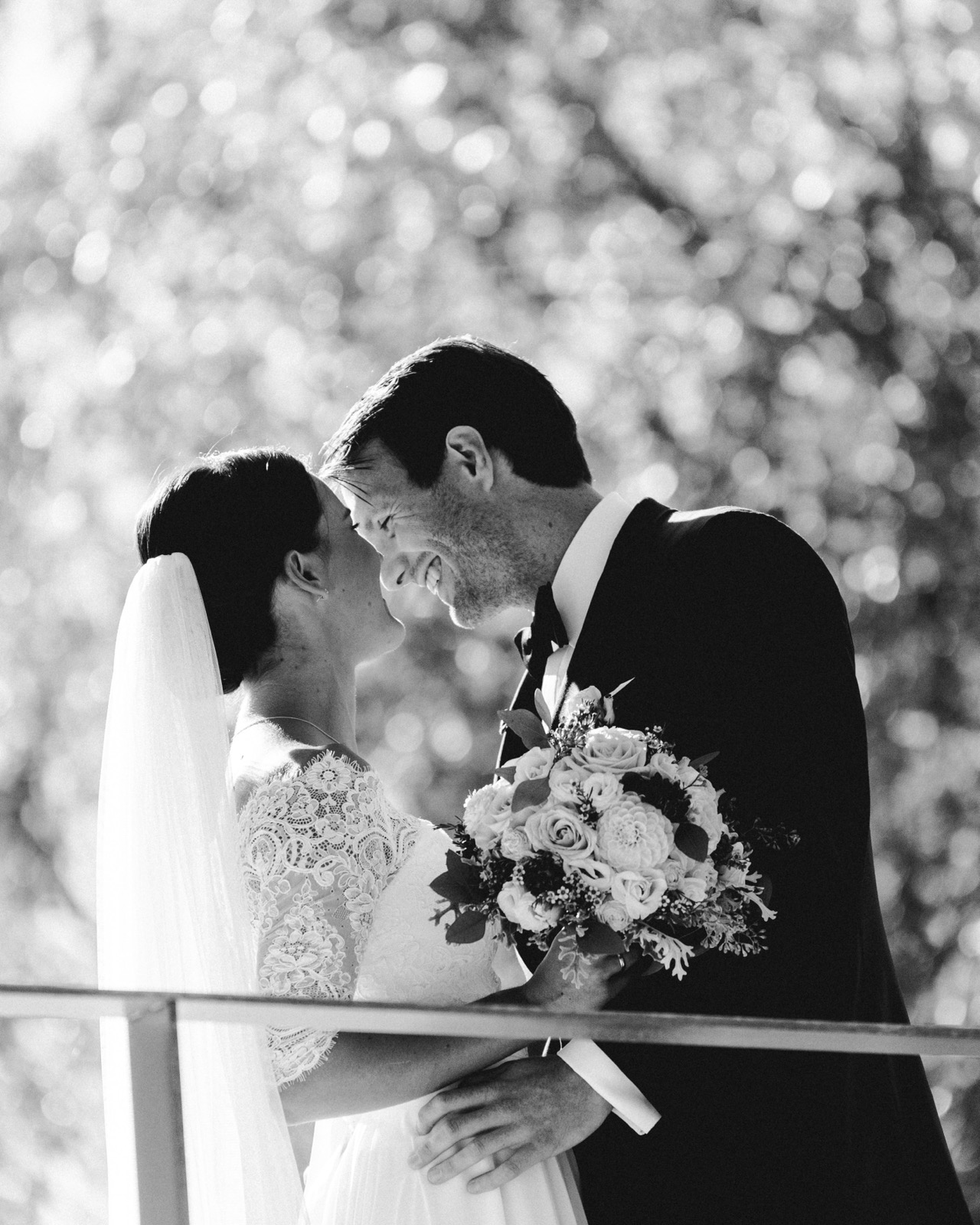 Schwarzweißes Porträt: Brautpaar lacht Stirn an Stirn, Bouquet im Vordergrund, Bokeh-Licht im Hintergrund