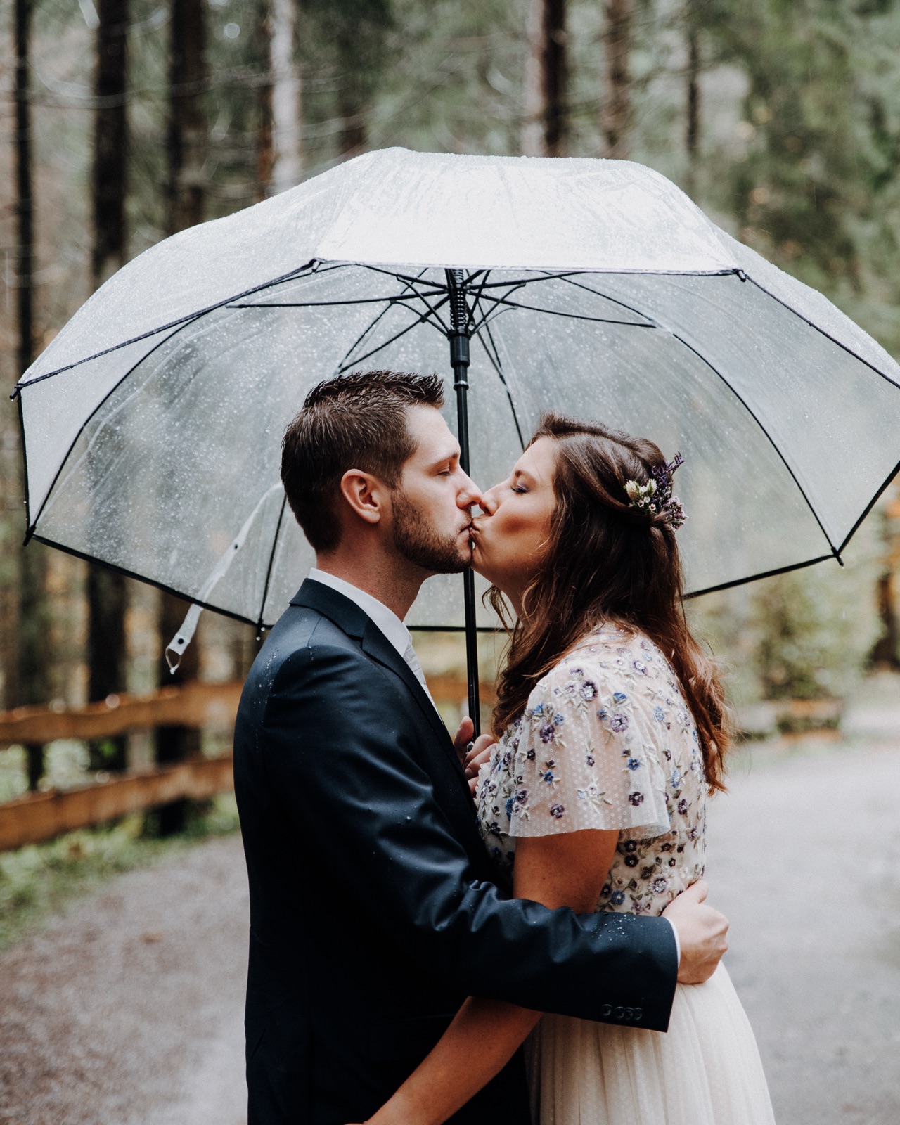 Brautpaar küsst sich unter einem durchsichtigen Regenschirm auf einem Waldweg, fotografiert von einem erfahrenen Hochzeitsfotografen