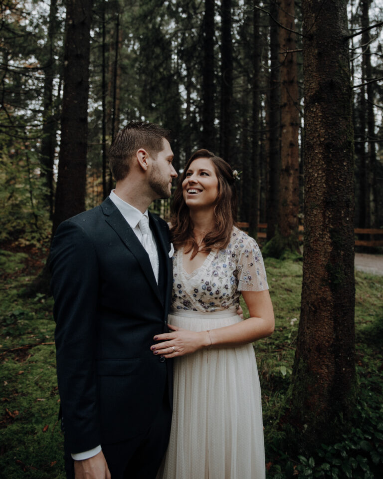 Brautpaar lacht sich im dunklen Tannenwald an, Braut im floral bestickten Kleid